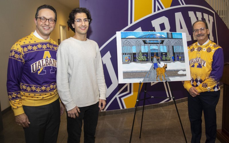 A student in a white shirt stands by a large image of his greeting card illustation, flanked by men wearing UAlbany shirts with snowflack designs