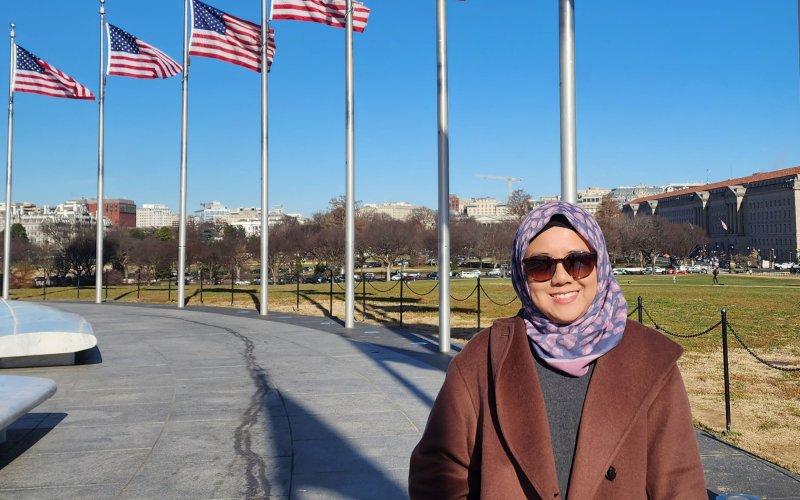 A smiling woman in a headscarf and winter coat stands on a path with flagpoles flying the American flag