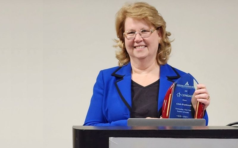 A woman with shoulder-length blonde hair and glasses in a blue blazer holds an award at a podium.