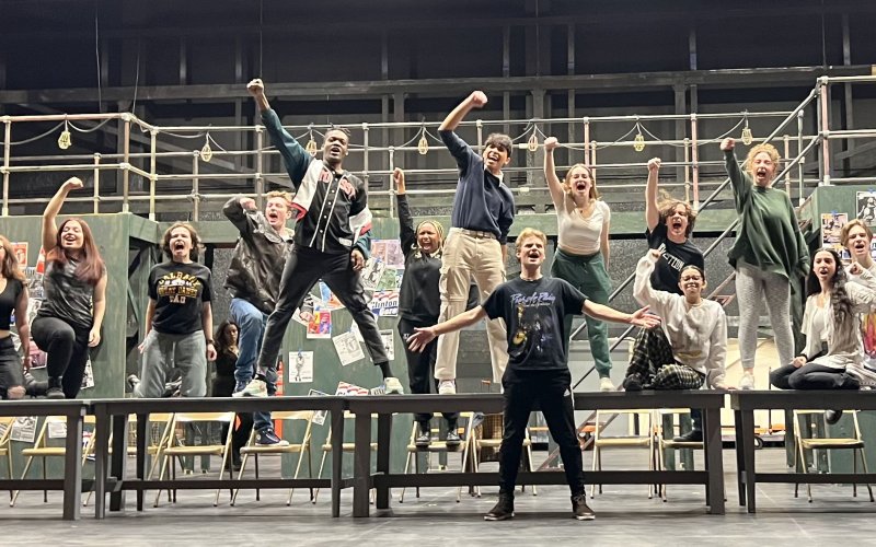 large cast of actors on platforms singing, most with one arm in the air