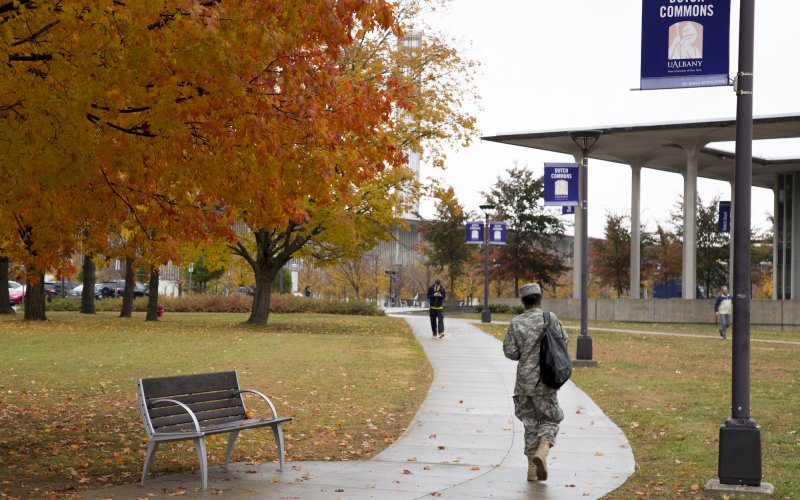 A person in a military uniform can be seen from behind walking along a path through Dutch Commons on the UAlbany campus on a wet fall day. A student walks toward them and trees display bright orange and yellow leaves.