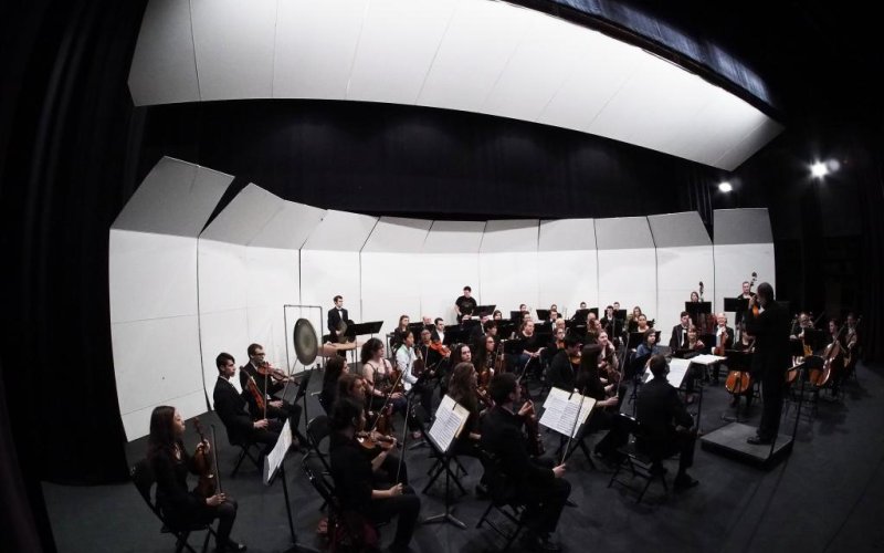 An orchestra wearing all black is pictured on a black stage surrounded by white lights and white acoustic panels.