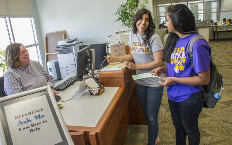 Two students wearing shirts that say UAlbany Great Danes stand in front of a desk where a woman seated behind the desk sits at a computer by a sign that says "Reference Ask Me I am Here to Help."