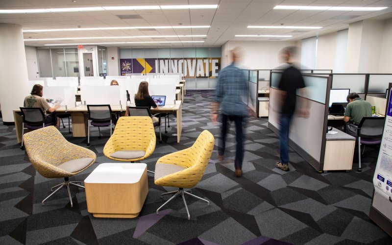an office with people seated at desks and others walking between cubicles with the word "Innovate" on a purple sign the window in white lettering.
