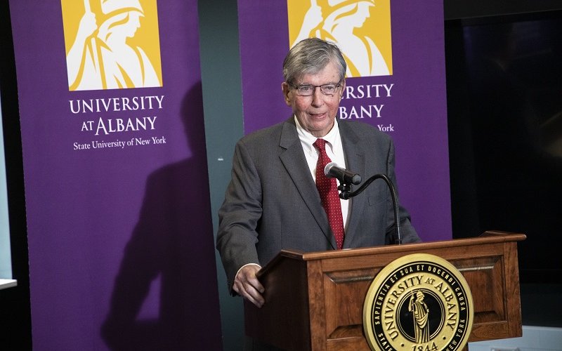 NYS Sen. Neil D. Breslin stands wearing a dark gray suit and red tie speaking at wooden lectern with the UAlbany seal framed on either side behind him by two purple UAlbany banners with the University's gold Minerva logo.