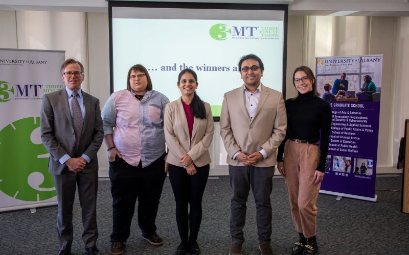 Four graduate students — the four finalists who presented at the live 3MT competition — stand with Vice Provost and Dean Kevin Williams in front of two banners, one featuring the Three Minute Thesis competition and the other advertising the Graduate School. A PowerPoint slide is in the background which says "and the winners are...".