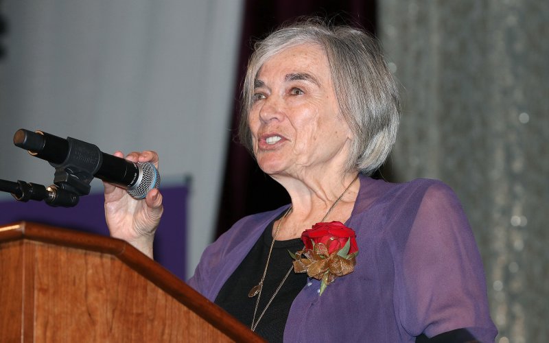 A woman with short gray hair wears a purple blouse and rose lapel as she stands at a podium with a hand grasping a microphone