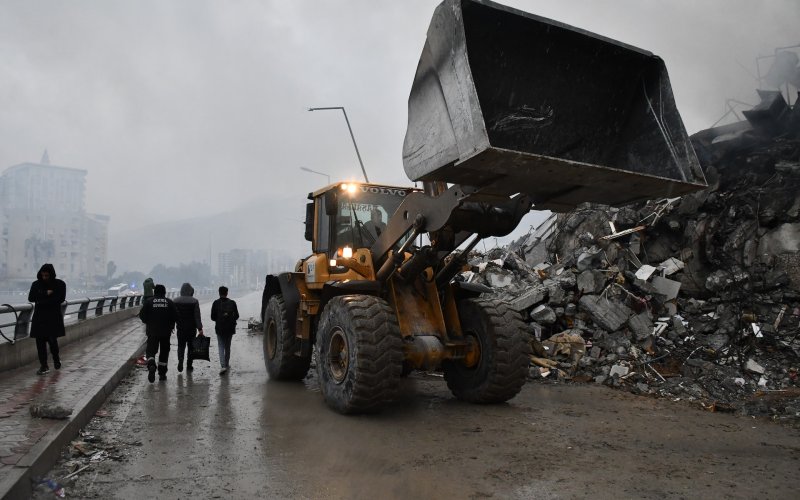 A bulldozer lifts its blade high into the air next to a pile of rubble along a pier as people dressed in black walk by. The sky is gray and hazy and the street is littered with debris.