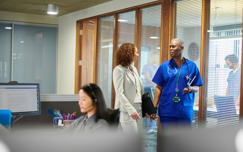 Three people - two standing and speaking to each other, one sitting at a computer wearing a headset - are pictured indoors in a healthcare setting.