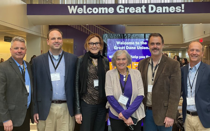 Six people in business attire and lanyards pose in the UAlbany Student Union. Behind them is a digital sign that reads "Welcome to the Great Dane Union", and "Welcome Great Danes" is painted on the wall above.