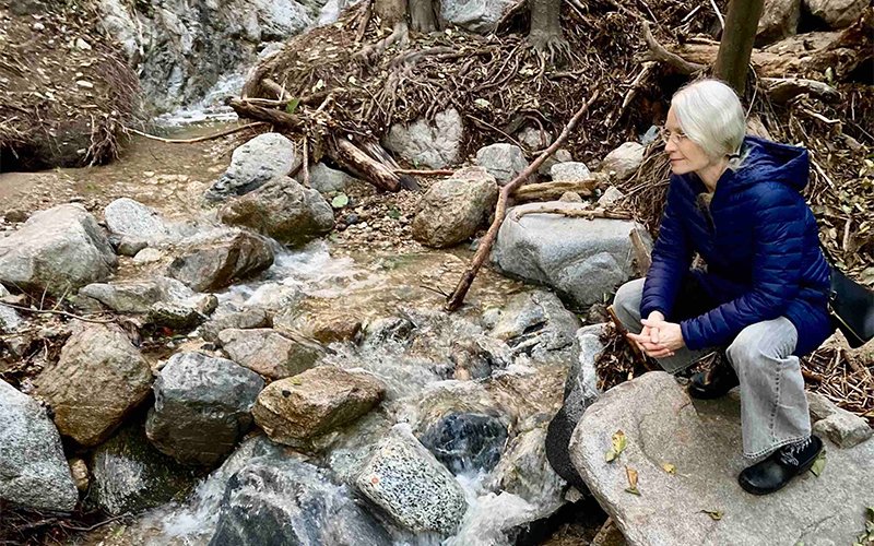 Laura Jones, with white hair, a blue jacket and jeans, squats and examines and flowing brook in Millard Canyon