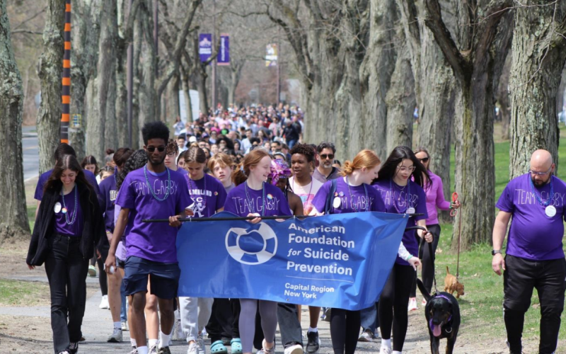 Students and community members gather to walk a course on UAlbany's Uptown Campus for the 2022 Out of the Darkness Walk. The day is sunny and a line of students leading the walking crowd hold a blue "American Foundation for Suicide Prevention" and wear purple T-shirts that say "Team Gabby". banner  
