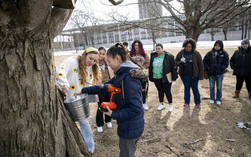 Olivia Dirla and Magnolia Roosa, from UAlbany's Office of Sustainability, demonstrate maple tree tapping for students at Dutch Quad.