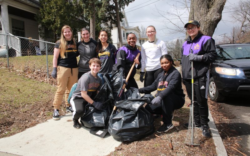 Eight people wearing UAlbany gear hold rakes and trash bags in an Albany neighborhood as they pose for a photo