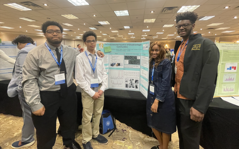 Four students — three boys and a girl wearing name tags and dress clothes — stand next to a poster board depicting air pollution research in a hotel conference room.
