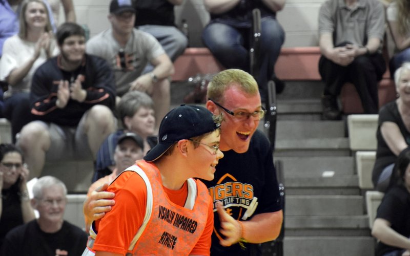 A student athlete celebrates with his visually impaired teammate after he scores in basketball as fans cheer from the stands.