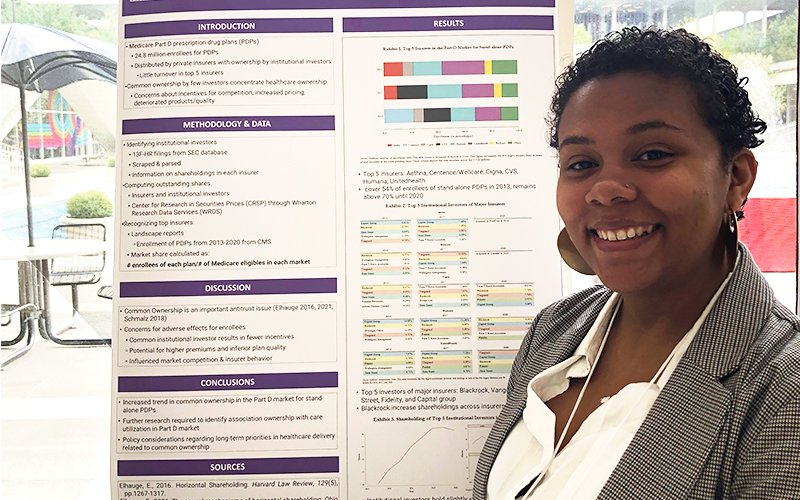 Alyssa Kamara smiles in front of a poster. On the poster, headings of white text in purple boxes organize the information. The headings read: "Introduction," "Methodology & Data," "Discussion," "Conclusions," "Sources," and "Results."