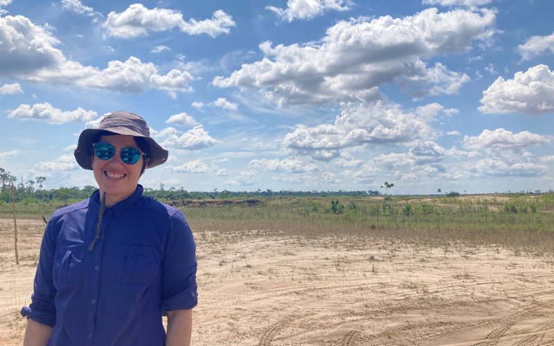 Beth Feingold stands at a former mining area in La Pampa, Madre de Dios, Peru. Feingold is wearing a blue button-down shirt, gray bucket hat and blue mirrored sunglasses. The background is mainly dry, cleared, open land, with forest in the far distance on the horizon. The sky is blue and full of large fluffy clouds. 