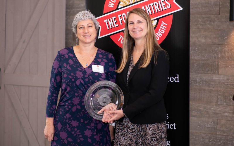 Janine and Natasha hold a circular glass award and stand in front of a sign that says The Food Pantries.