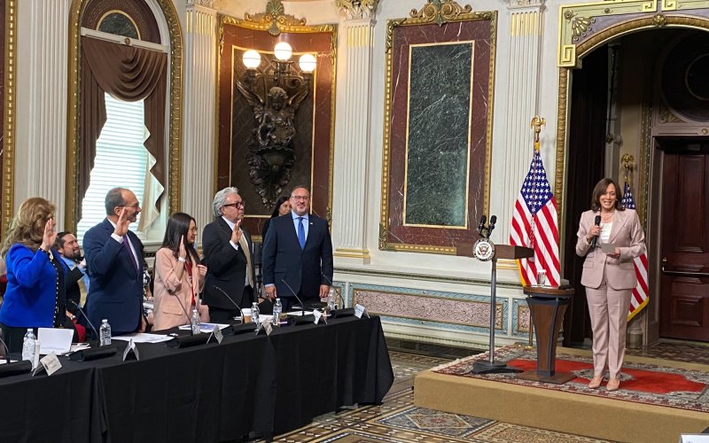 Five people stand at a long table with their right hands raised as they are sworn in by Vice President Kamala Harris, who is standing on a riser at the front of an ornate room in front of to U.S. flags.