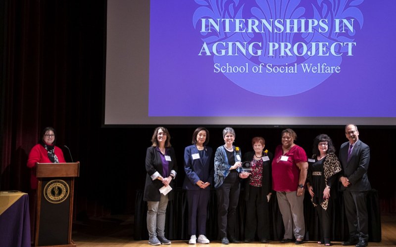 A woman stands at a podium near a line of seven smiling people standing under a screen with the words "Internships in Aging Project/ School of Social Welfare"
