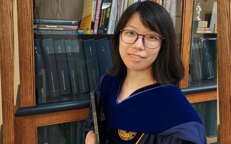A women wearing glasses and a blue velvet PhD hood stands in front of a bookcase