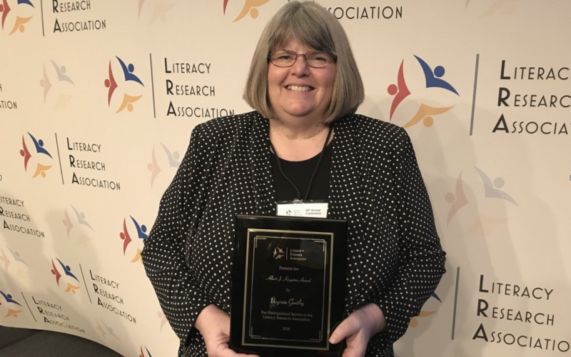 A woman with chin-length gray hair and glasses wears a polka dot blazer and holds a plaque. The wall behind her has repeating logos next to the words “Literacy Research Association.”