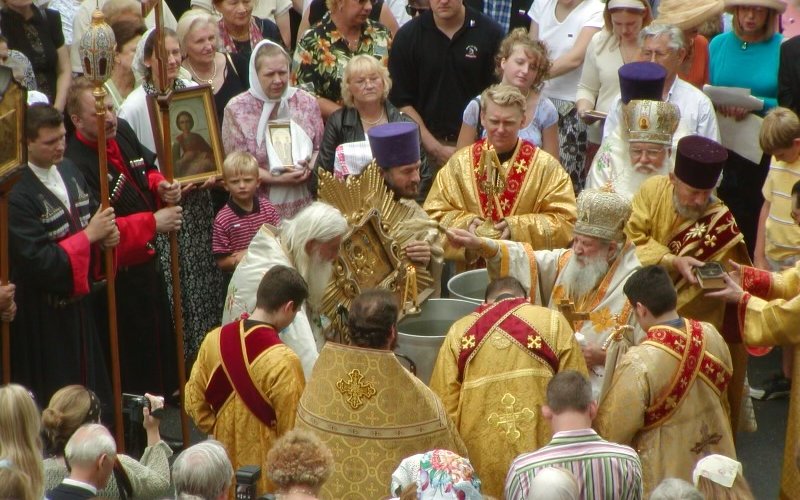 Orthodox Christians in gold ceremonial garb participate in the blessing of the water rite at St. Vladimir Memorial Church in Jackson, N.J. A crowd watches, some holding framed pictures.