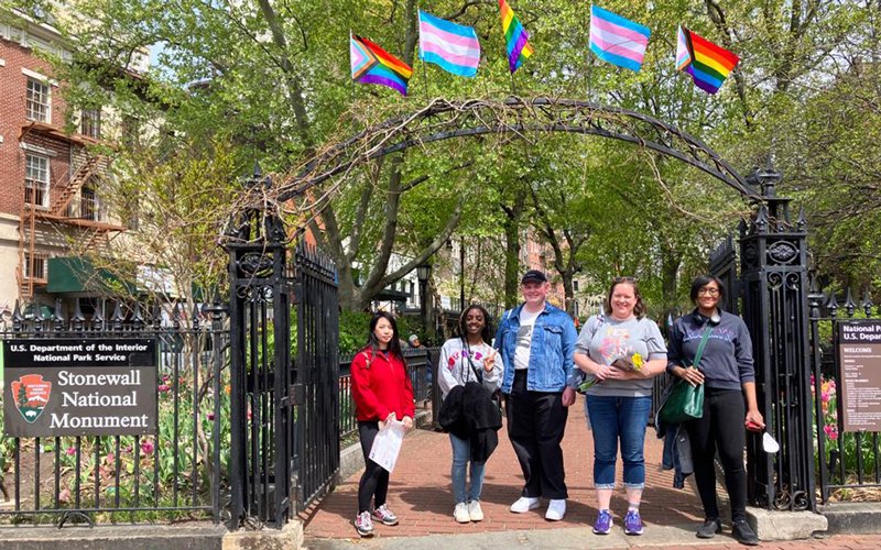 Five students stand underneath the archway of the Stonewall National Monument gate. Above them fly the traditional pride flag, pride progress flags and the transgender pride flag. A sign to the left reads, "U.S. Department of the Interior National Parks Service Stonewall National Monument."
