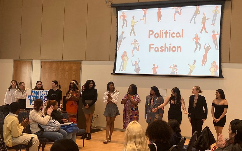 Thirteen women stand in front of a crowd dressed in a variety of outfits, one holding a sign that reads "Protect Reproductive Rights." Behind them, "Political Fashion" is displayed on the projector.
