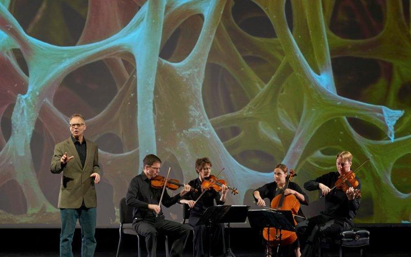 four musicians with instruments and a speaker in front of a projected image of DNA
