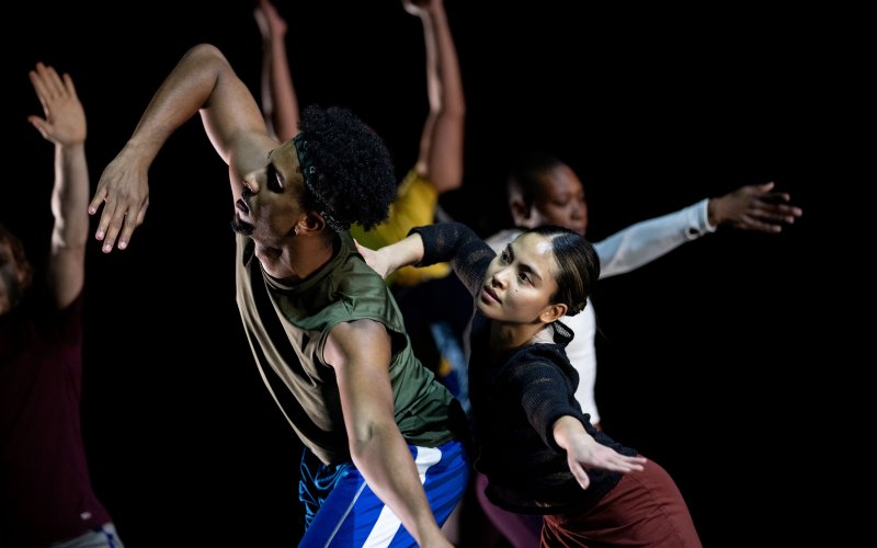 two dancers in foreground lean sideways in connection to one another; arms of other dancers are visible in background 