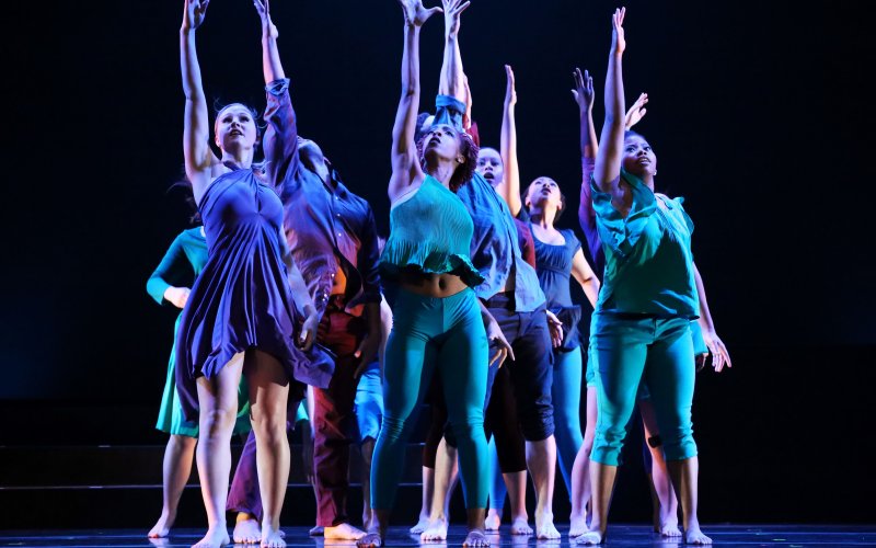 group of dancers in blue and green costumes stand in a clump with one arm raised in the air