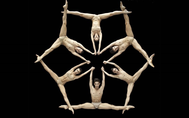 an overhead shot of dancers in nude costumes shows them connected by hands with arms overhead and legs in splits with ankles overlapping