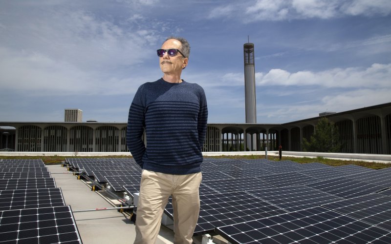 A man with short gray hair and sunglasses poses next to solar panels on the UAlbany campus on a blue-sky day.