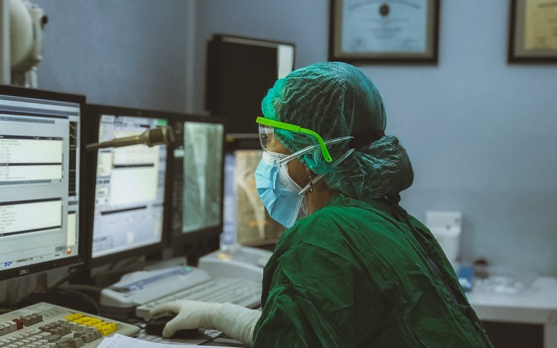 A healthcare worker wearing scrubs, goggles, mask and gloves, works at bank of computers in a dimly-lit room. 