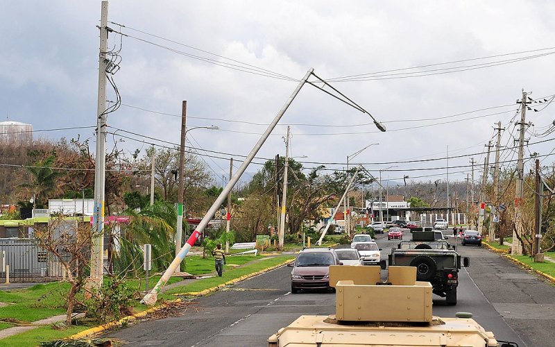 A convoy of military vehicles drive down a road in Puerto Rico with damaged power lines and light poles.