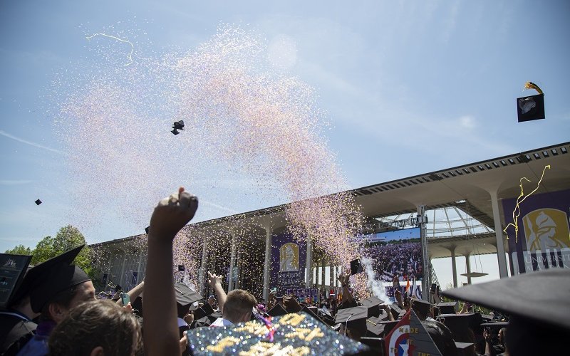 A confetti cannon shoots a blast of purple and gold confetti over a sea of black mortar board hats and hands raised in celebration at UAlbany's spring 2023 Undergraduate Commencement.
