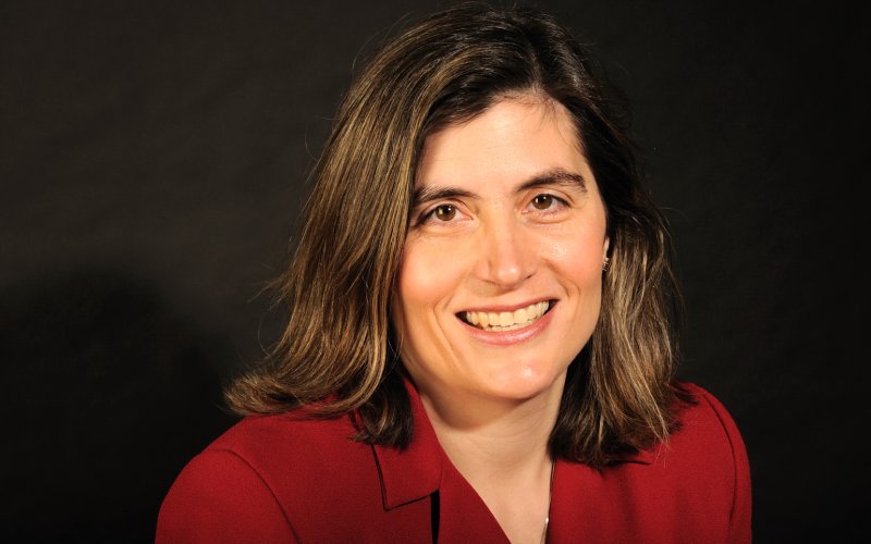 Portrait of Erin Bell; Erin is smiling wearing a red blazer with a dark brown backdrop