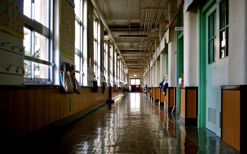 An empty hallway of a school building, with light reflecting off the floor and tote bags hanging on hooks on the walls