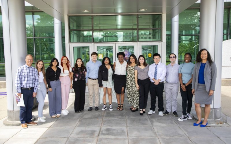 Twelve undergraduate students stand in a row in front of the Life Science building entranceway. RNA Institute director Andy Berglund is standing on the far left. Shanise Kent, assistant dean of Graduate Education, is standing on the far right. It is a sunny day, but the group is standing in the shade of an awning attached to the building entrance. Most of the building behind them is glass, with silver colored columns and trim.