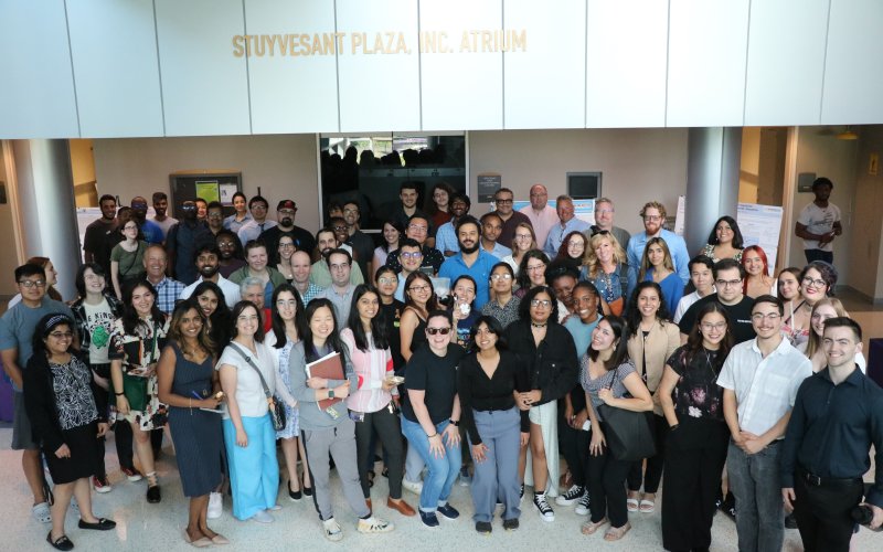 A large group of smiling students and researchers are gathered in a light filled atrium in the RNA Institute.