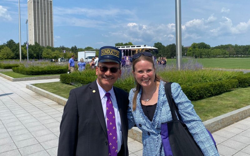 President Havidán Rodríguez and Jessica Castner, both smiling broadly, pose together in front of UAlbany's new electric trolley. It is a beautiful, sunny summer day. The background landscape is full of green grass, full green trees, and planters with boxwood bushes and purple flowers.