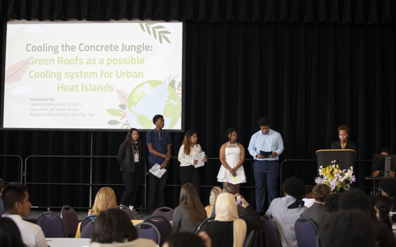 Six students stand on a stage next to a podium and microphone and in front of a large screen titled "Cooling the Concrete Jungle: Green Roofs as a possible Cooling system for Urban Heat Islands." A seated audience looks on.