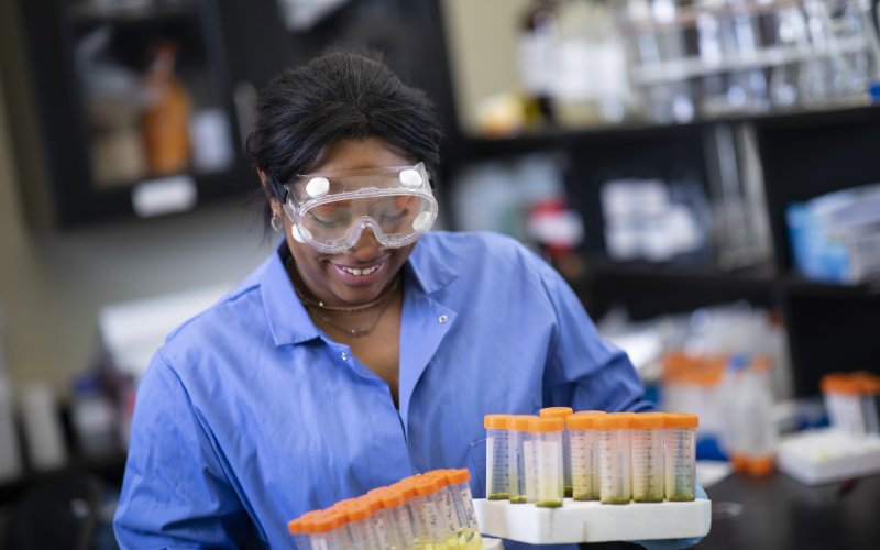 A woman wearing safety goggles and a blue shirt smiles as she holds a case of vials in each hand inside a laboratory space.