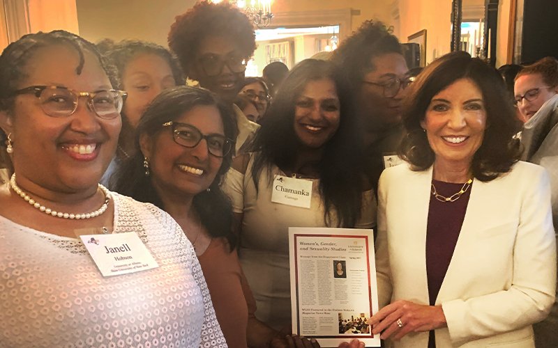 Governor Kathy Hochul holds the WGSS newsletter while surrounded by smiling WGSS faculty and students in a hallway of the Governor's Mansion.