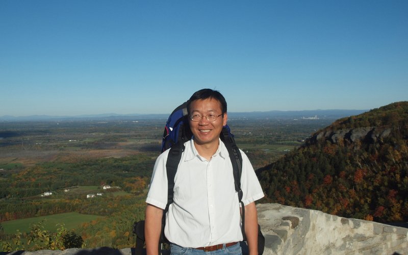 Professor Rongwei Yang stands at Thacher Park, in front of a view full of trees, fields and changes leaves