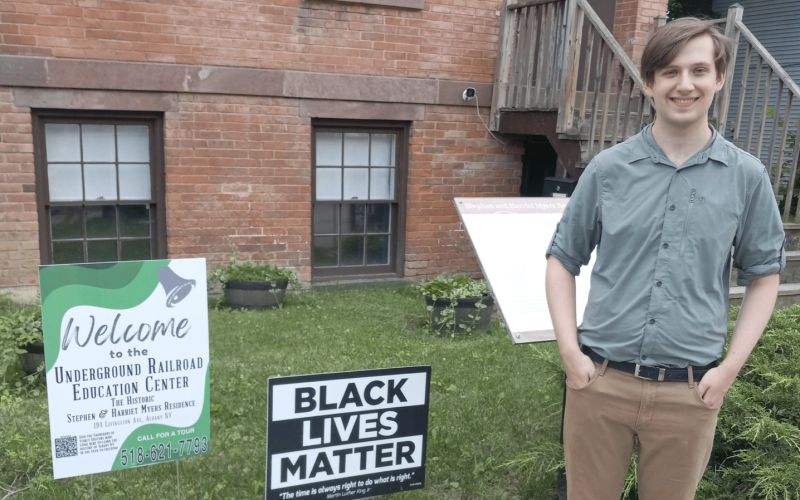 A young man with light brown hair in a button down shirt and tan pants stands in front of a brick building next to a "Black Lives Matter" sign and sign that reads "Welcome to the Underground Railroad Education Center."