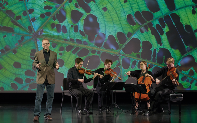 A man in a black button down, jeans and blazer speaks on a stage. Nearby a string quartet dressed in all black plays, bows poised, in front of a large screen depicting a close-up image of the scales of a leaf.