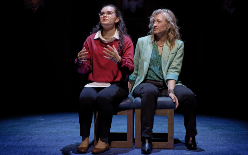 Two women are seated next to each other on a stage. The woman on the left, in a red sweater over a white button down shirt, gestures with her hands while staring in the distance as the other woman, in a green blouse and blazer, watches her.